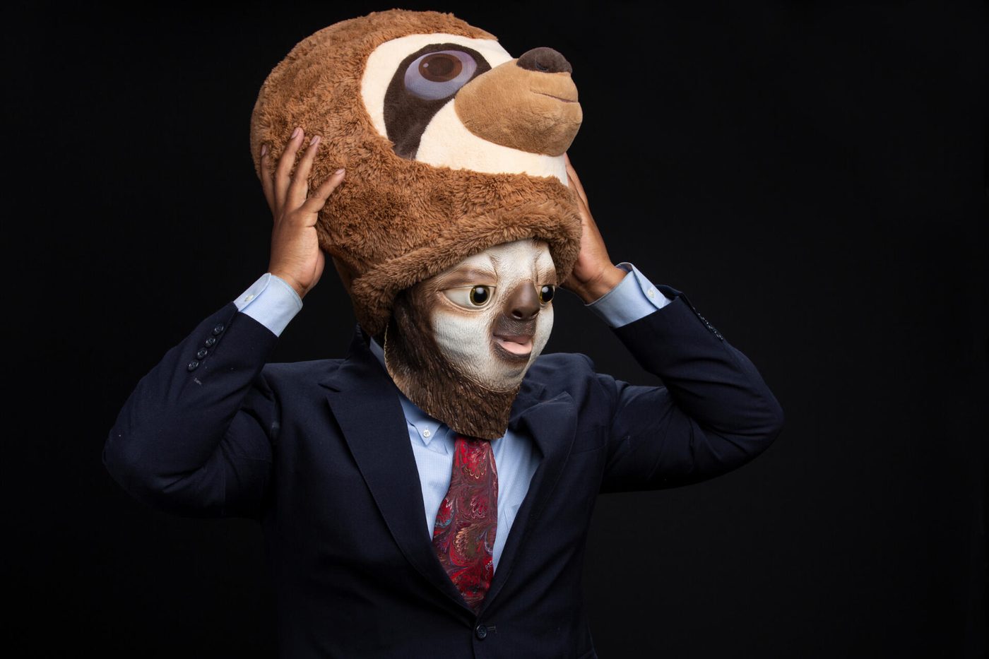 Ferris the Sloth: a man in a suit and red patterned tie lifting a plush sloth mask onto his head against a black backdrop